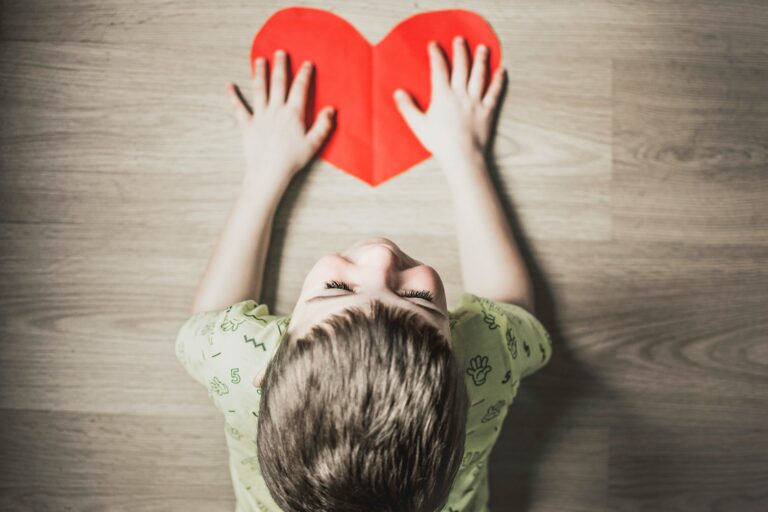A child holds a red paper heart on a light wooden floor