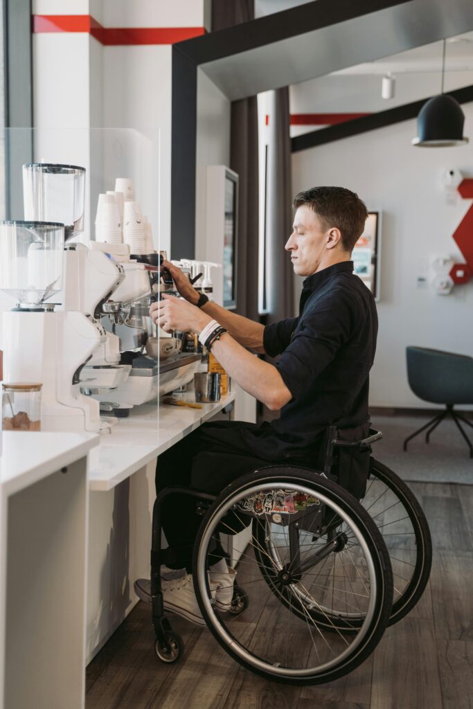 A wheelchair using man makes coffee at an accessible station in a brightly lit room