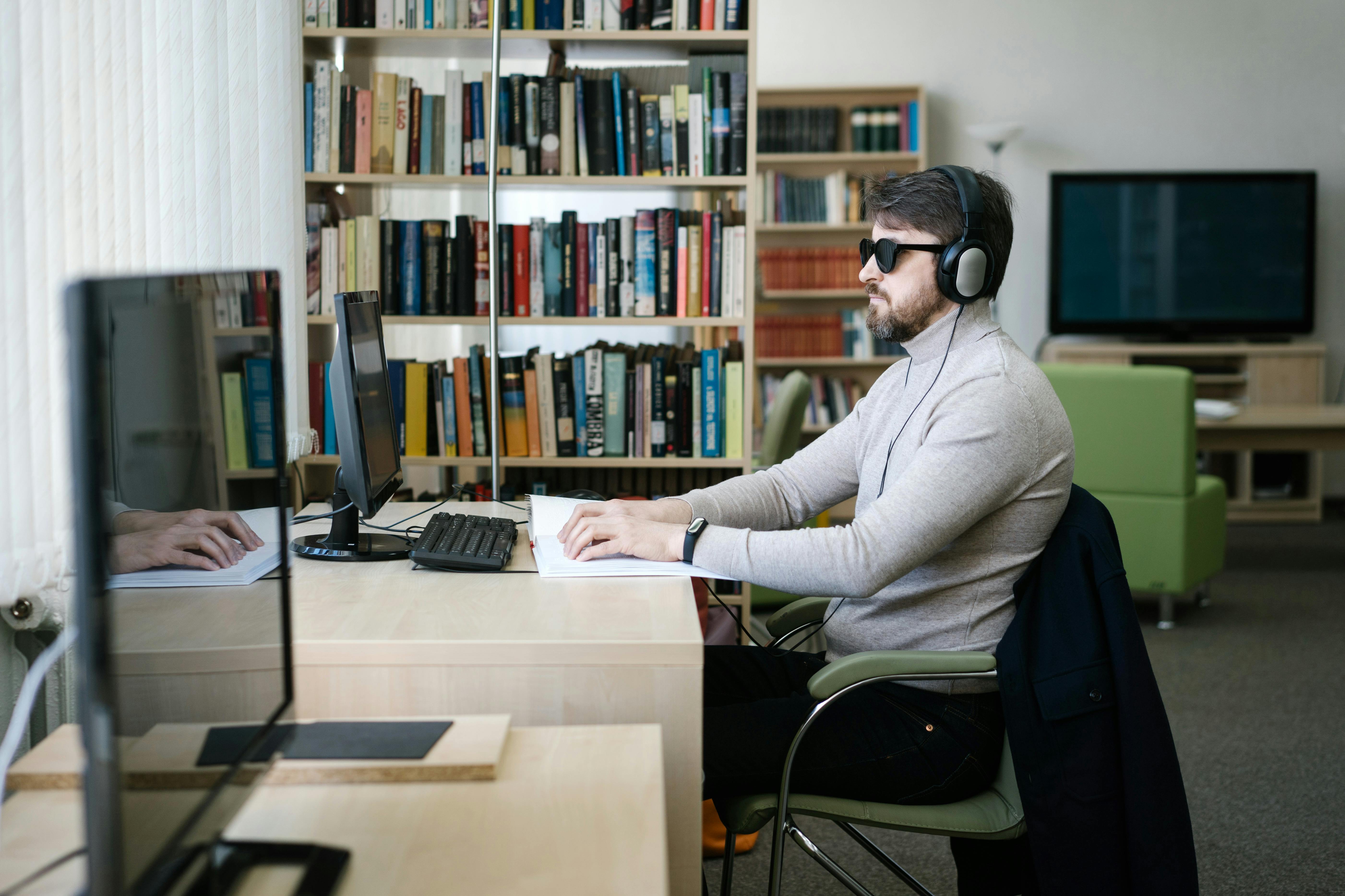 A man sits at a desk by a bright window with dark glasses and headphone, whilst reading braille with his hands